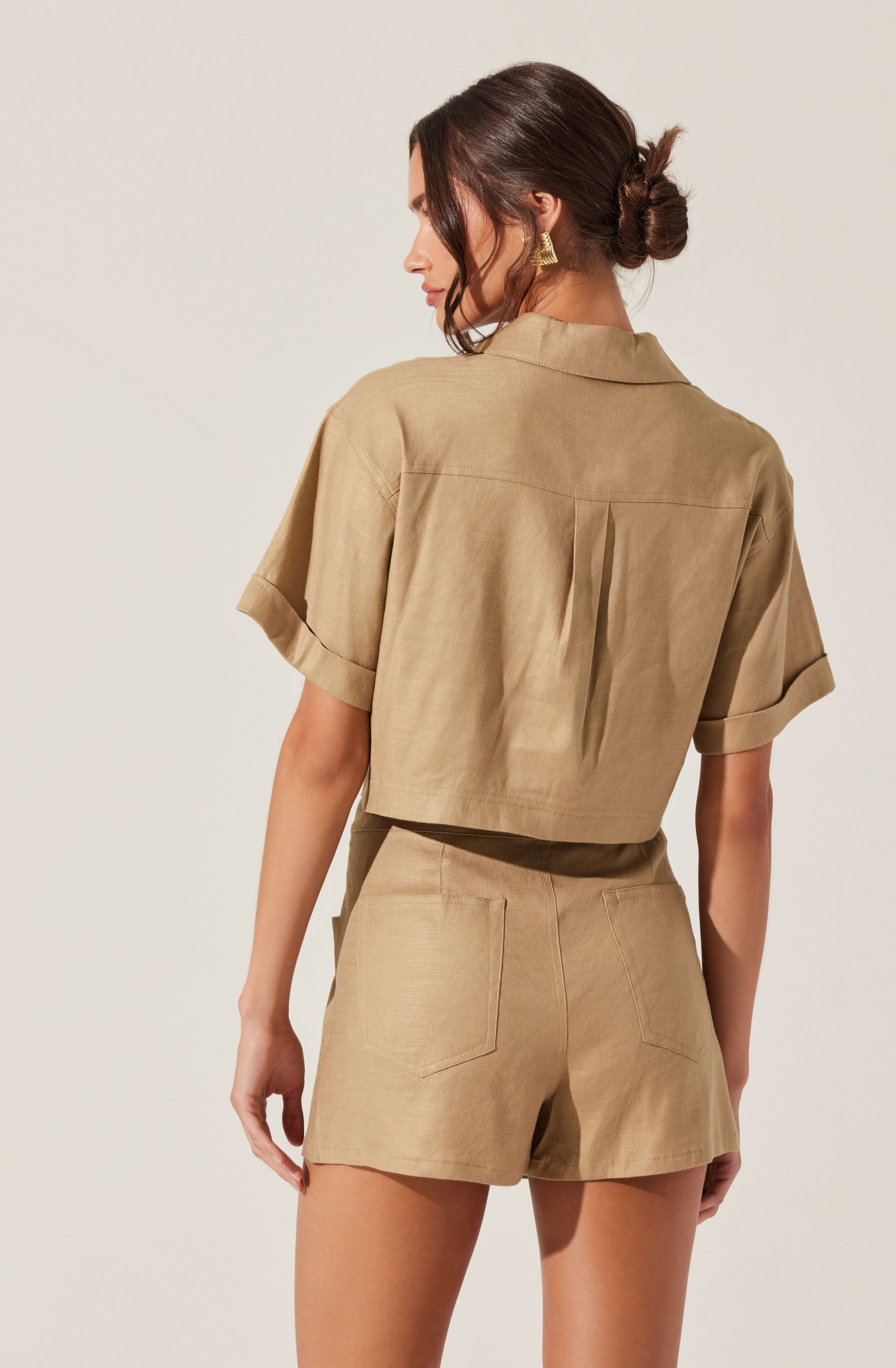 A woman is wearing a tan, short-sleeved, cropped button-up shirt with a collar and pleats in the back, paired with matching high-waisted shorts that have two back pockets.