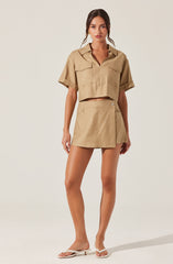 A woman is wearing a beige, short-sleeved, collared shirt with two large chest pockets. She pairs it with a matching beige mini skirt that has a side button detail. She is also wearing white sandals with a low heel.