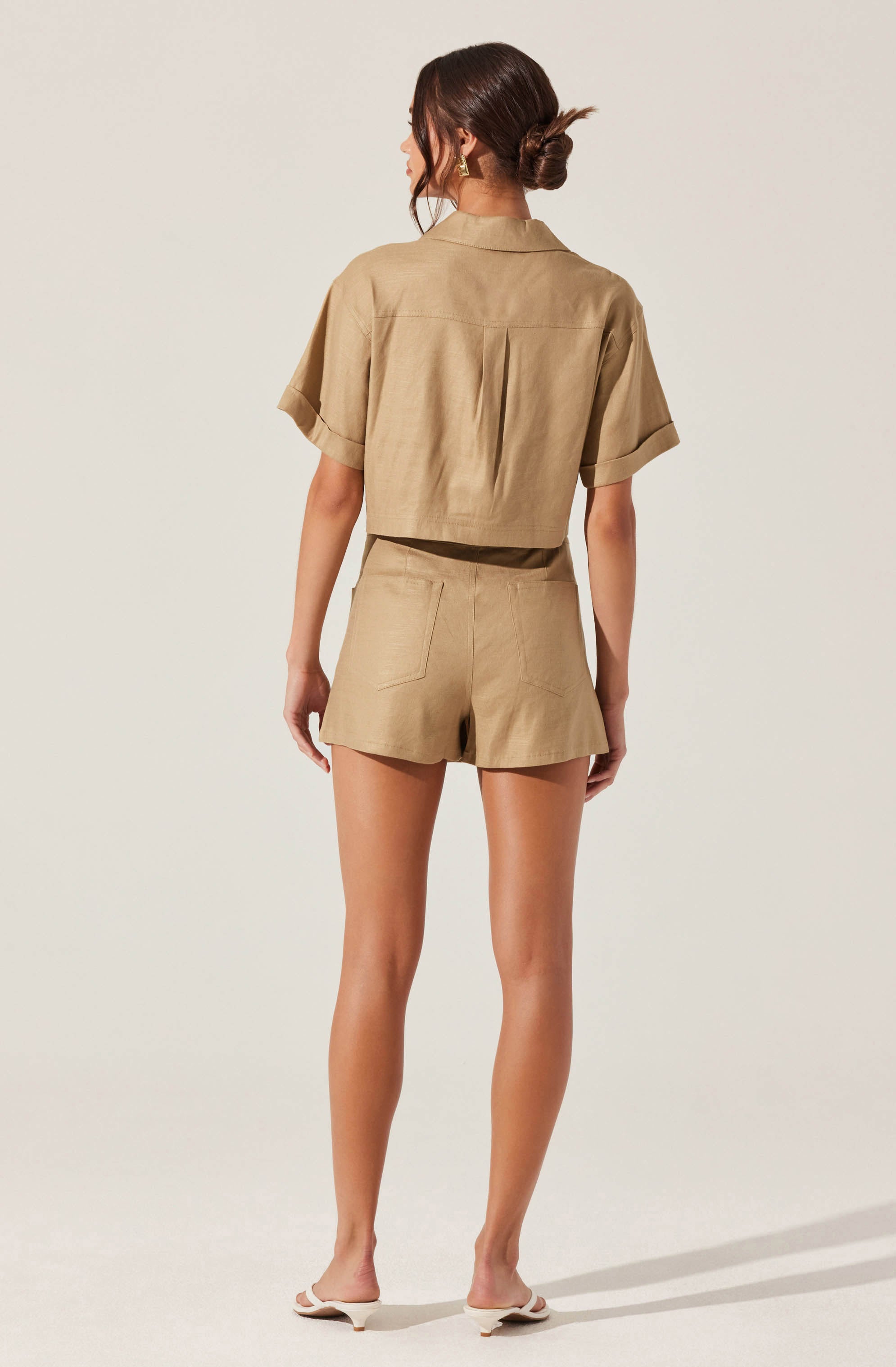 A woman is wearing a matching beige short-sleeved shirt and high-waisted shorts set. The shirt has a loose fit, a collar, and rolled-up sleeves. The shorts have a relaxed fit with visible back pockets. She is also wearing white heeled sandals.