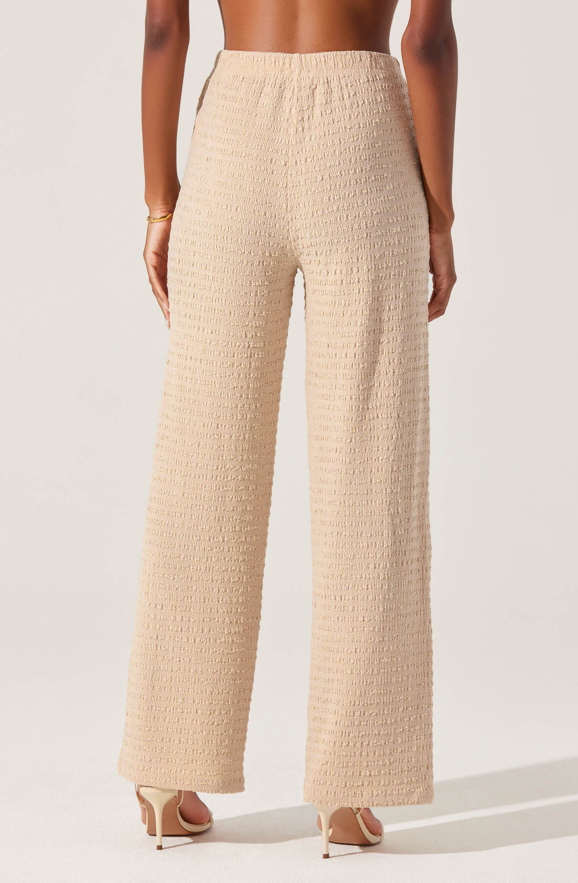 A person is wearing wide-leg, high-waisted trousers made of textured, cream-colored fabric with a subtle grid pattern. They are also wearing nude high-heeled shoes.