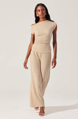 A woman is wearing a matching beige outfit consisting of a textured sleeveless top with a one-shoulder design and wide-leg pants with a similar textured pattern. She has long, dark hair and is accessorized with gold earrings, a gold bracelet, and rings. She is standing against a plain, light background.
