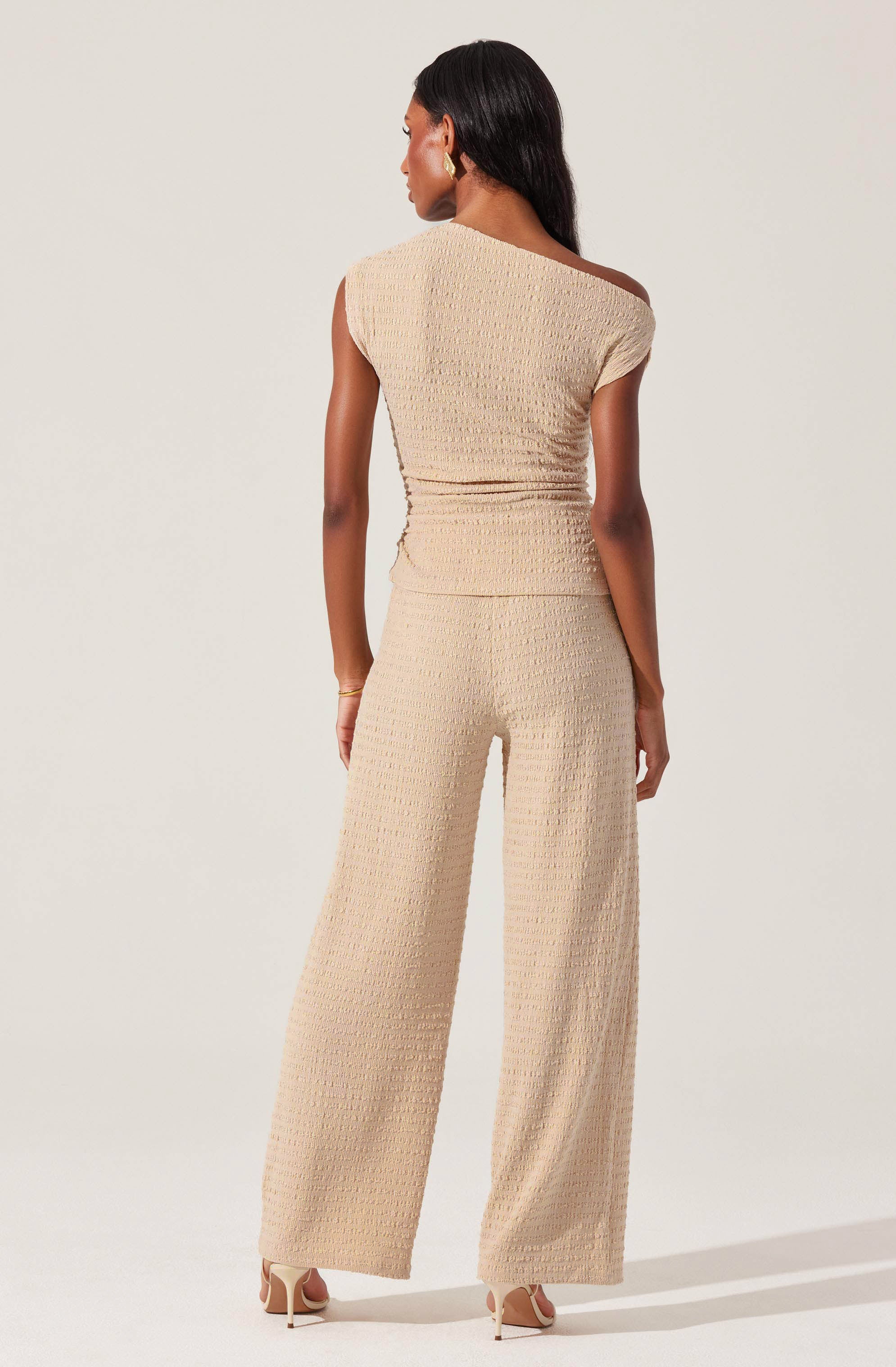 The woman is wearing a beige sleeveless top with a textured, waffle-like pattern and matching wide-leg pants made of the same textured fabric. The outfit appears elegant and comfortable, with the top having a cap sleeve on one side and a unique shoulder detail. She is accessorized with gold earrings and a gold bracelet.