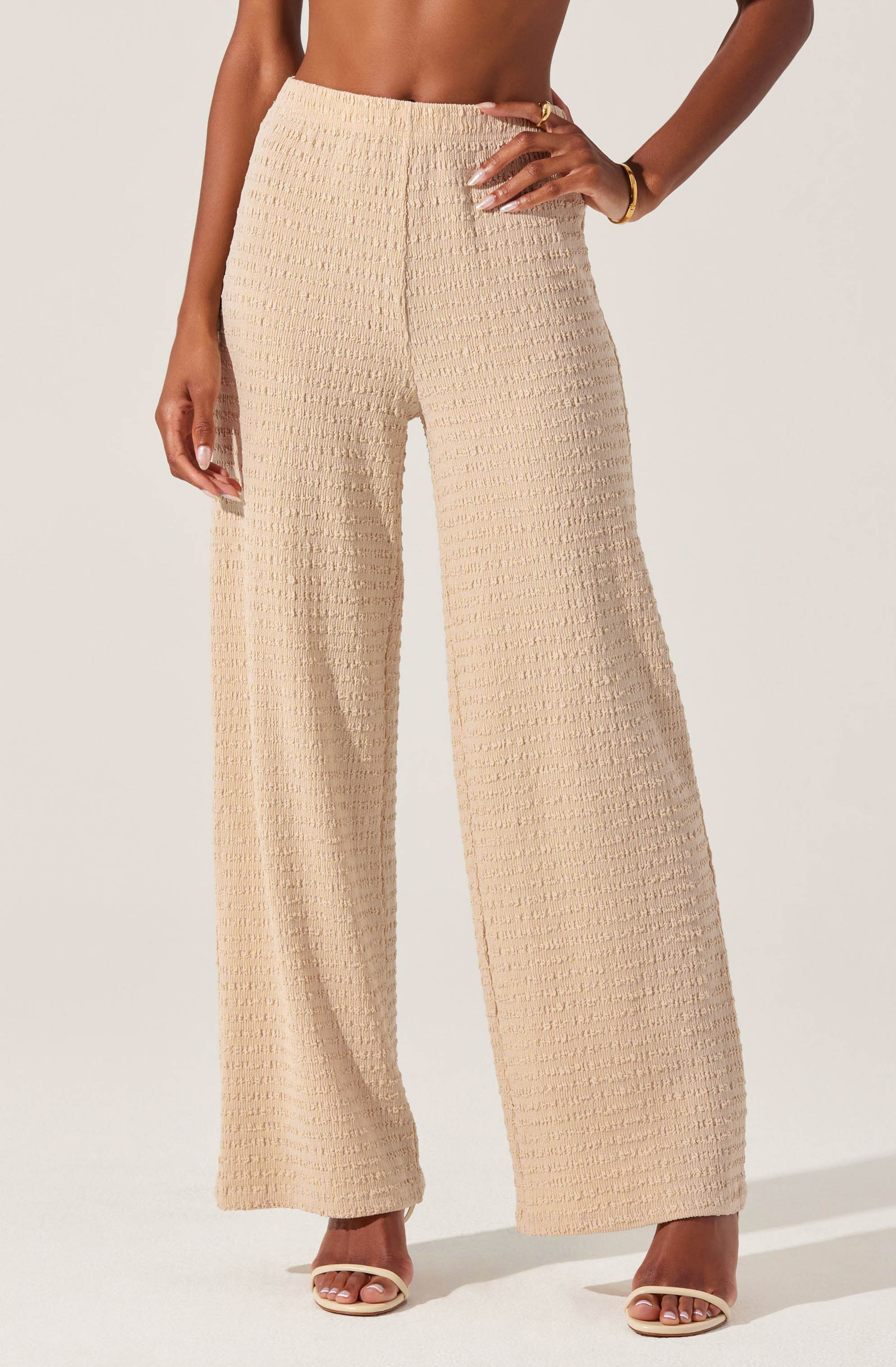 A person is wearing high-waisted, wide-leg beige pants made of textured fabric with a subtle grid pattern. They are paired with open-toe sandals with a thin strap across the toes and a thicker strap around the ankle. The person's right hand rests on their hip, and they have a gold bracelet on their right wrist.