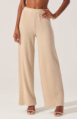 A person is wearing high-waisted, wide-leg beige pants made of textured fabric with a subtle grid pattern. They are paired with open-toe sandals with a thin strap across the toes and a thicker strap around the ankle. The person's right hand rests on their hip, and they have a gold bracelet on their right wrist.
