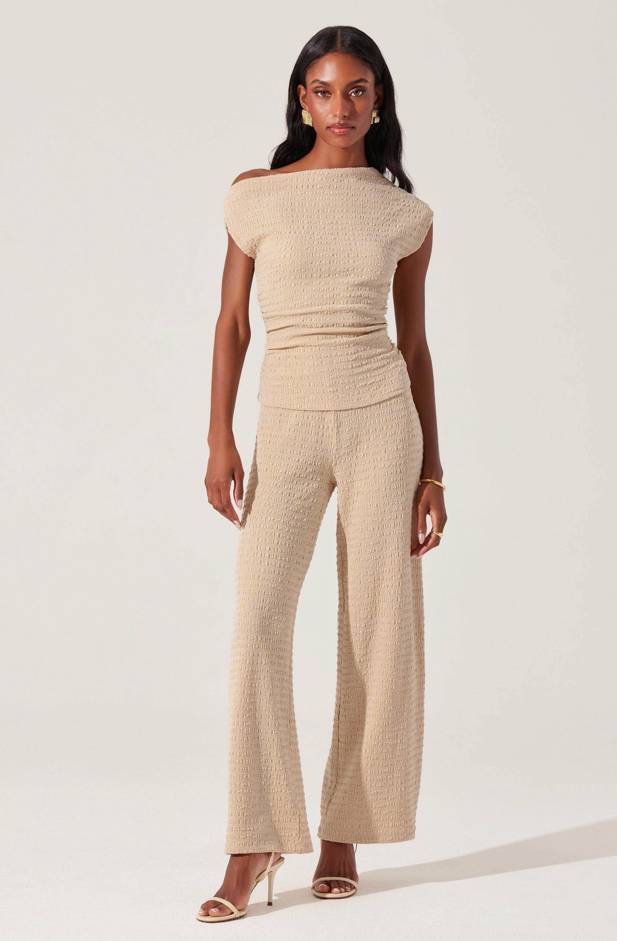 A woman is wearing a matching beige outfit consisting of a textured, short-sleeved top with a wide boat neckline that falls slightly off her shoulders, and high-waisted, wide-leg pants made from a similar textured fabric. She has accessorized with gold earrings, a gold bracelet, and is wearing nude-colored high-heeled sandals.