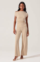 A woman is wearing a matching beige outfit consisting of a textured, short-sleeved top with a wide boat neckline that falls slightly off her shoulders, and high-waisted, wide-leg pants made from a similar textured fabric. She has accessorized with gold earrings, a gold bracelet, and is wearing nude-colored high-heeled sandals.