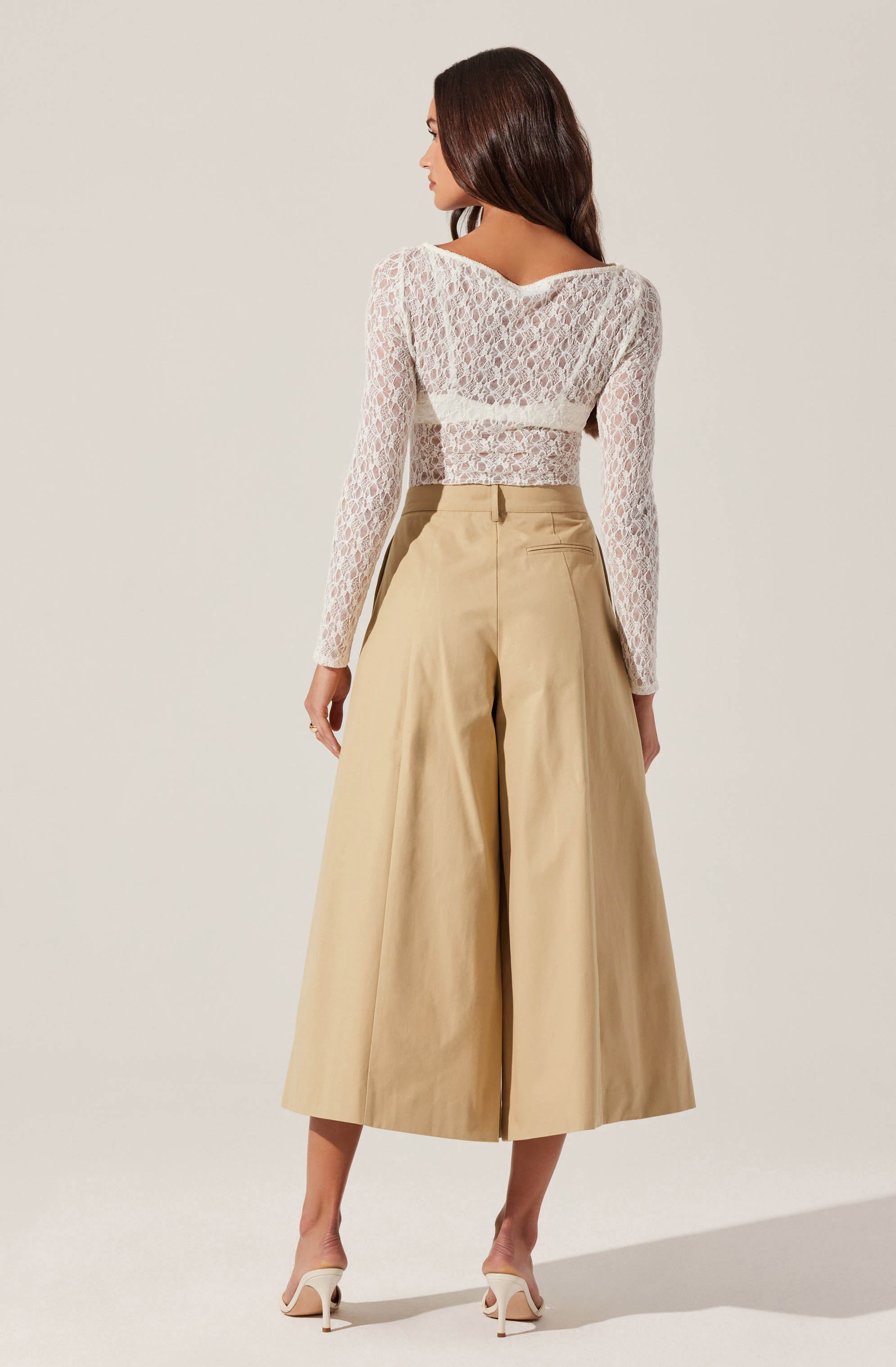 The woman is wearing a long-sleeved, semi-sheer white lace top with a floral pattern that covers her shoulders and arms. She pairs it with high-waisted, wide-leg beige cropped trousers that reach mid-calf. She completes the outfit with light-colored high-heeled sandals.