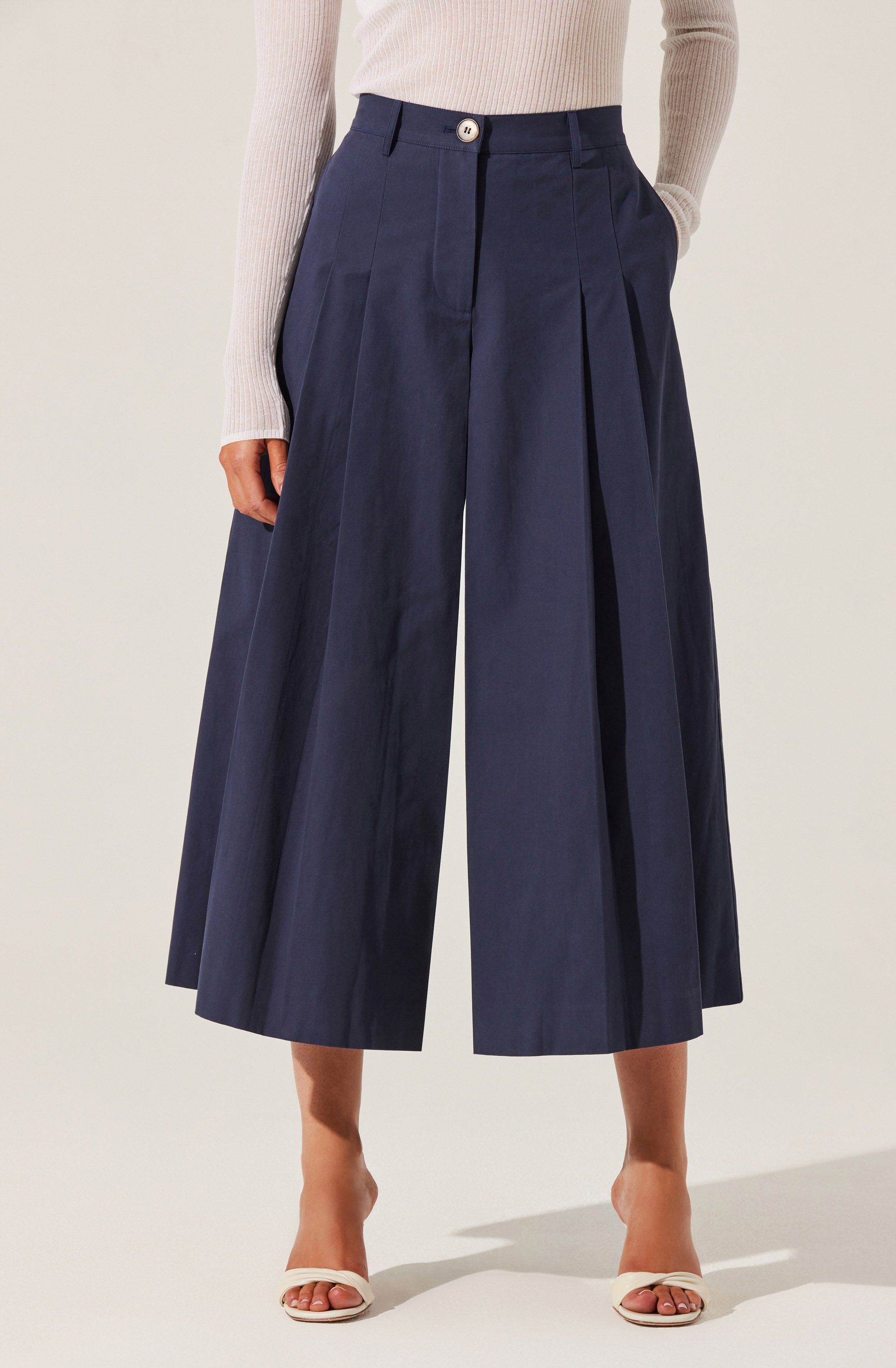A person is wearing high-waisted, wide-leg navy blue culotte pants with pleats in the front. The pants have a button closure at the waist and belt loops. They are paired with a light-colored, ribbed long-sleeve top tucked into the pants and open-toe white heels.