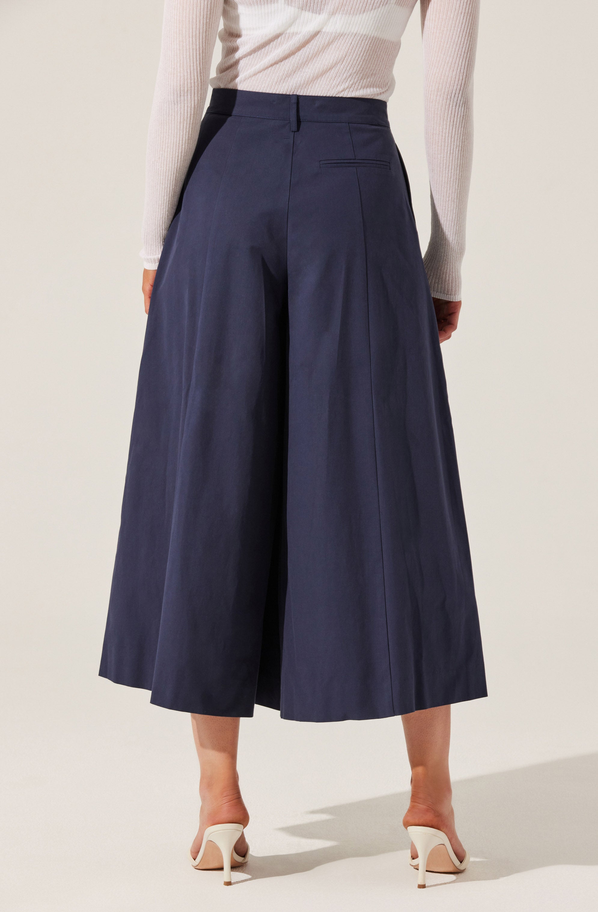 A woman is wearing high-waisted, wide-leg navy blue culottes that reach mid-calf. The culottes have a tailored fit with visible seam details and a back pocket. She pairs the pants with a long-sleeved, light-colored, ribbed top and white high-heeled shoes.