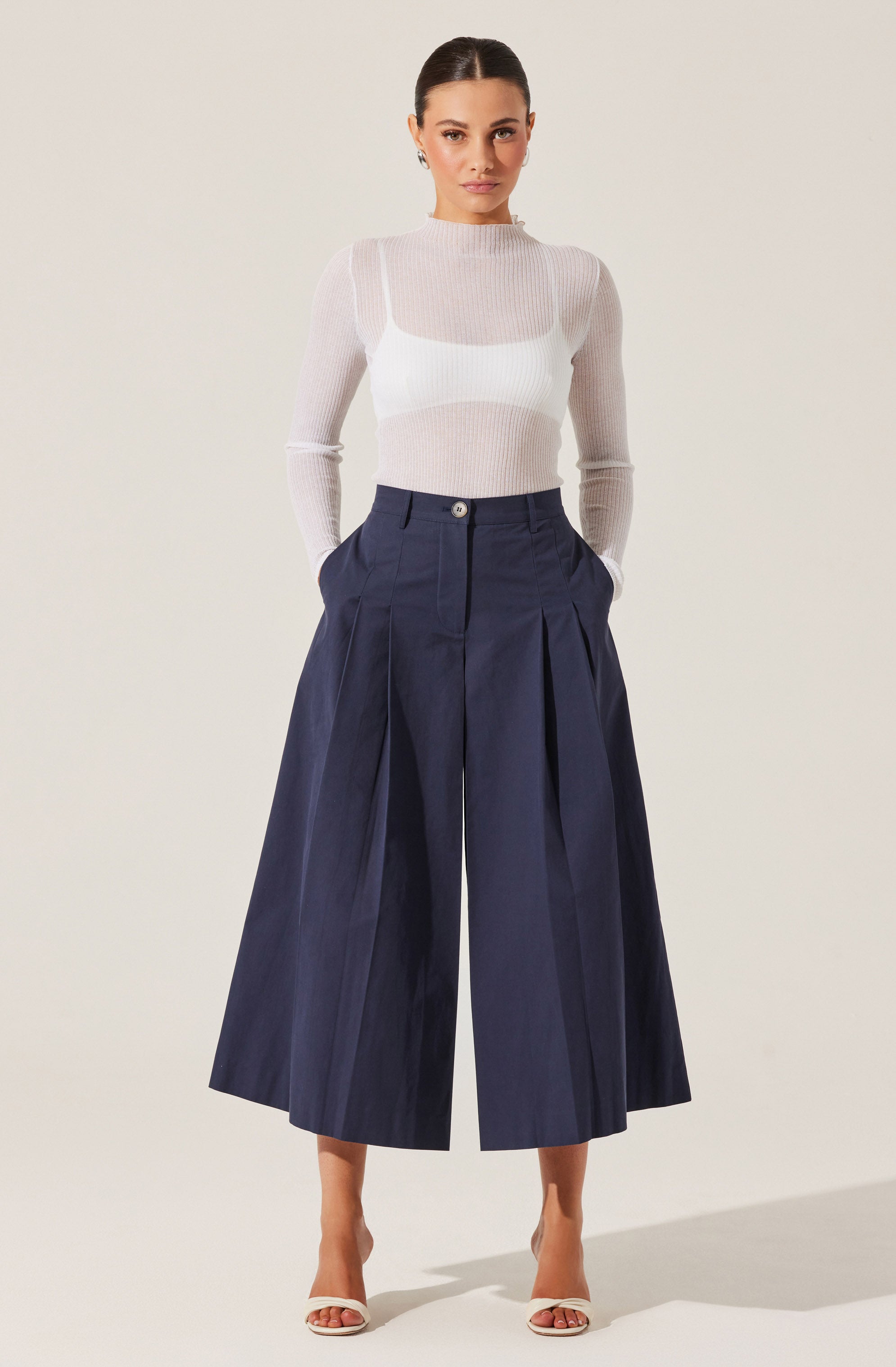 A woman is wearing a fitted, long-sleeved, light beige or cream-colored sheer knit top with a high neckline. She pairs it with high-waisted, wide-leg navy blue culotte pants that feature front pleats and a button closure at the waist. She completes the outfit with a pair of open-toe heeled sandals.