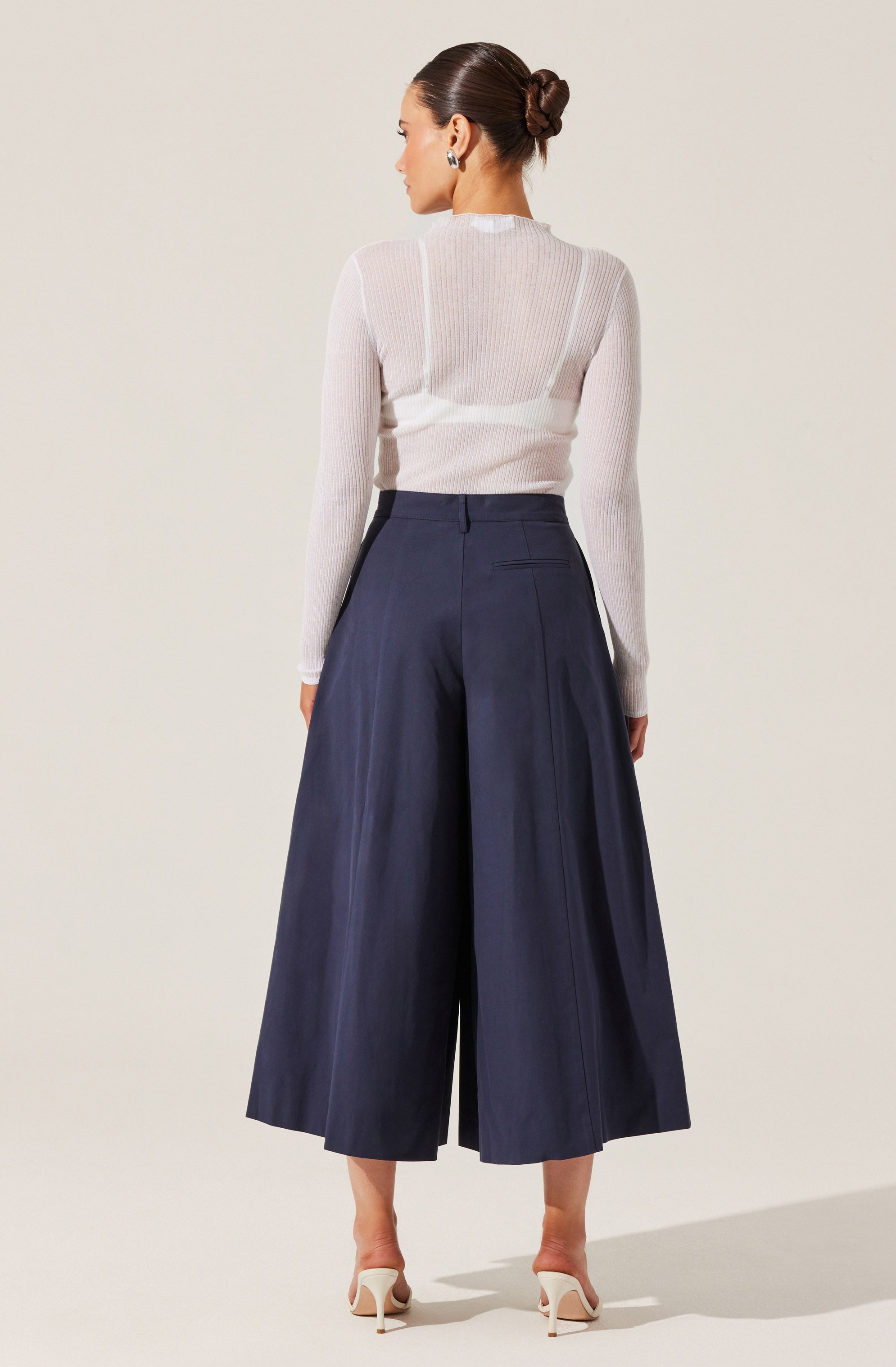 A woman is wearing a sheer, long-sleeved white top with a round neckline tucked into high-waisted, wide-leg navy blue culottes that reach mid-calf. She is also wearing white high-heeled shoes and has her hair styled in a bun.