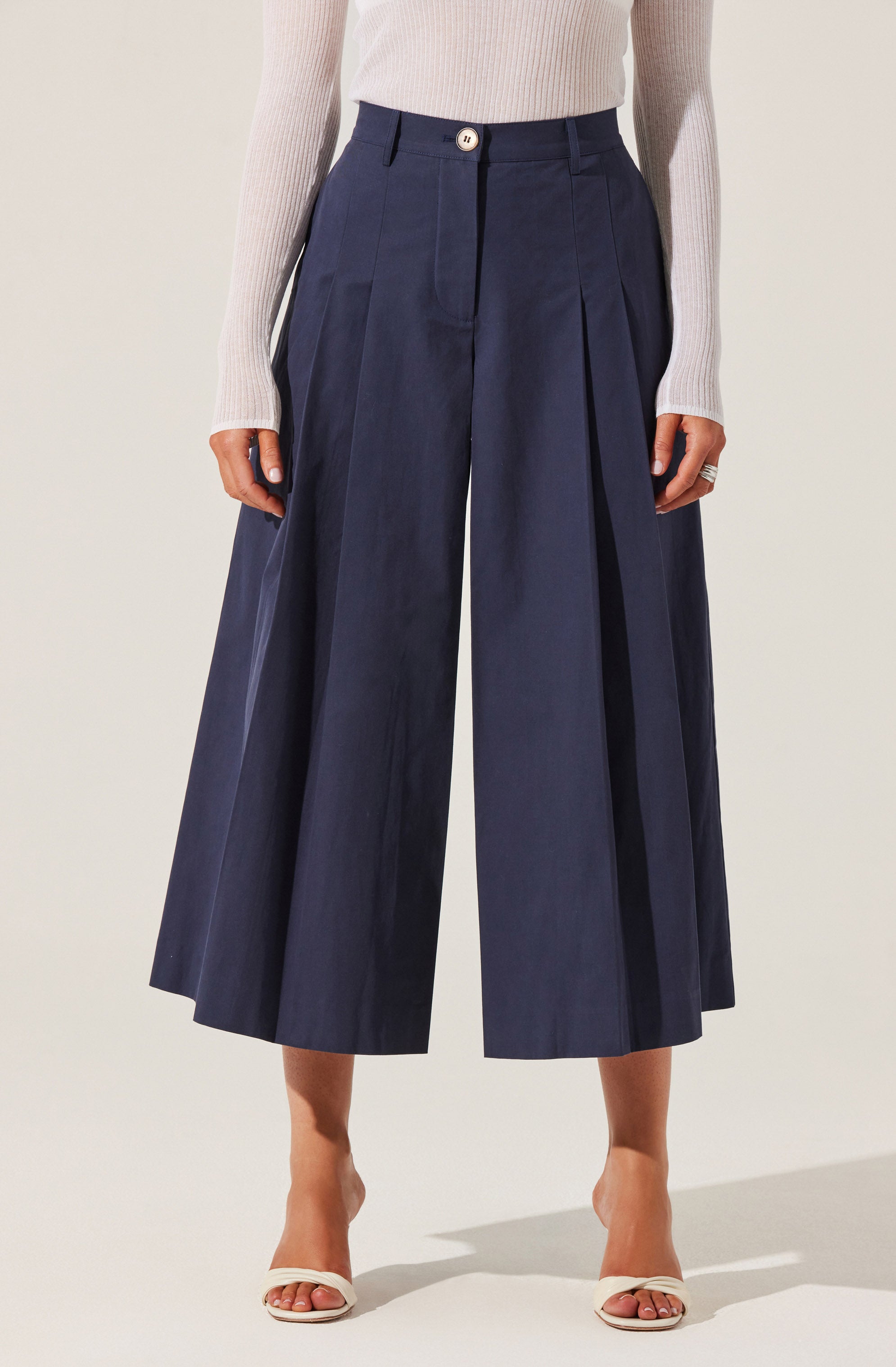 The image shows a person wearing wide-leg, high-waisted navy pants with a pleated design. The pants have belt loops and a white button at the waistband. The individual is also wearing a light-colored, long-sleeve ribbed sweater and open-toe nude high heels.
