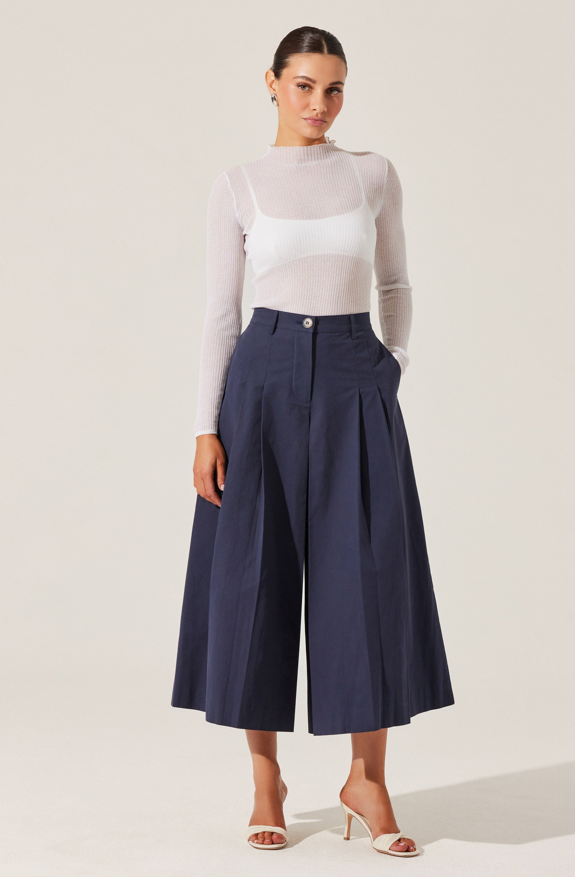 A woman is wearing a light, semi-sheer, long-sleeve knitted sweater in a neutral color, paired with high-waisted, wide-leg navy blue culottes that feature pleats and a single visible button at the waistband. She completes the outfit with light-colored open-toe heels.