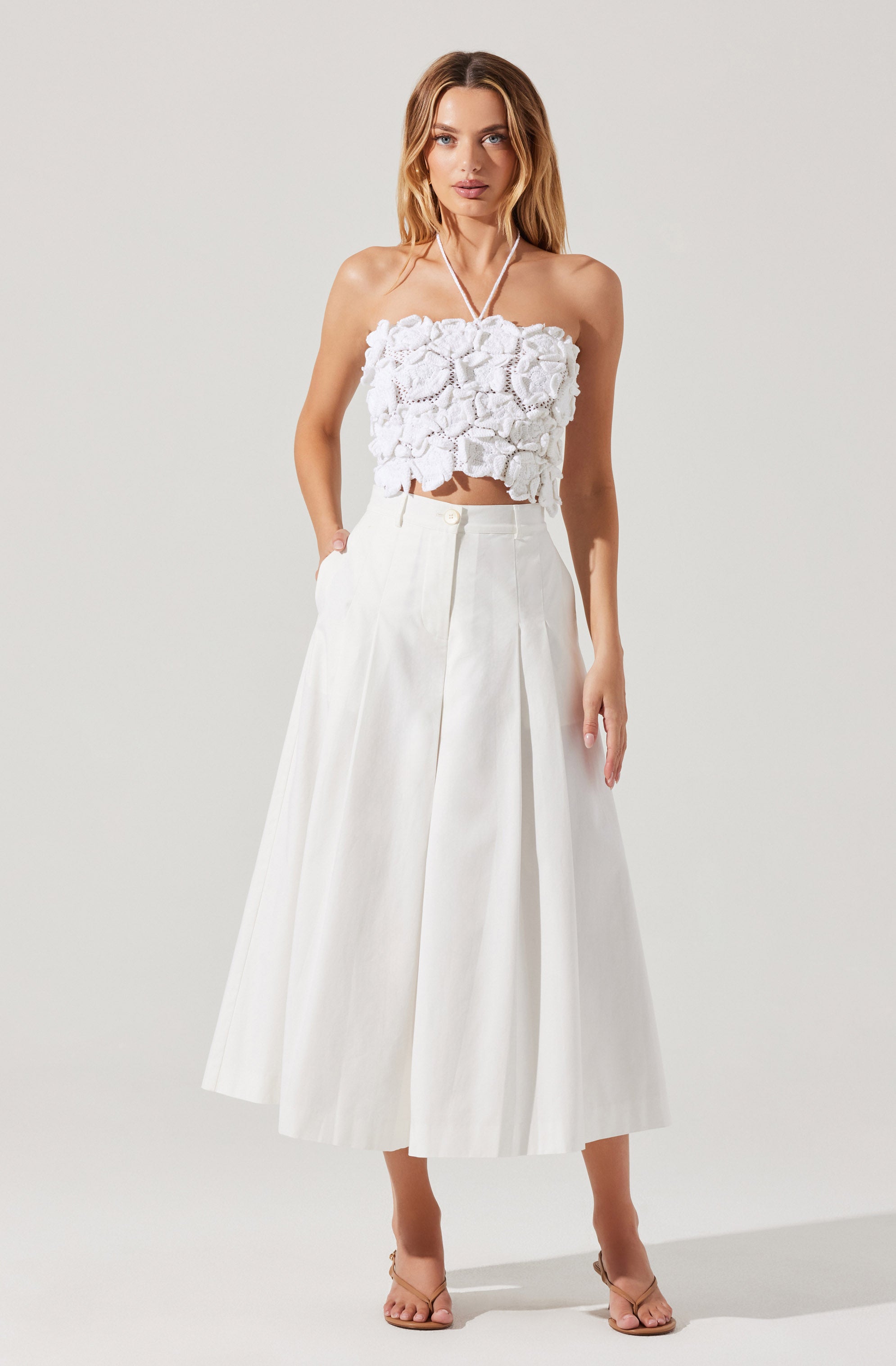 A woman is wearing a white sleeveless cropped top with textured, three-dimensional floral appliqué details. She pairs it with a high-waisted, wide-leg midi skirt in white, which features pleats and a subtle A-line silhouette. She completes the outfit with nude-colored strappy heeled sandals.