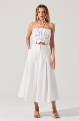 A woman is wearing a white sleeveless cropped top with textured, three-dimensional floral appliqué details. She pairs it with a high-waisted, wide-leg midi skirt in white, which features pleats and a subtle A-line silhouette. She completes the outfit with nude-colored strappy heeled sandals.