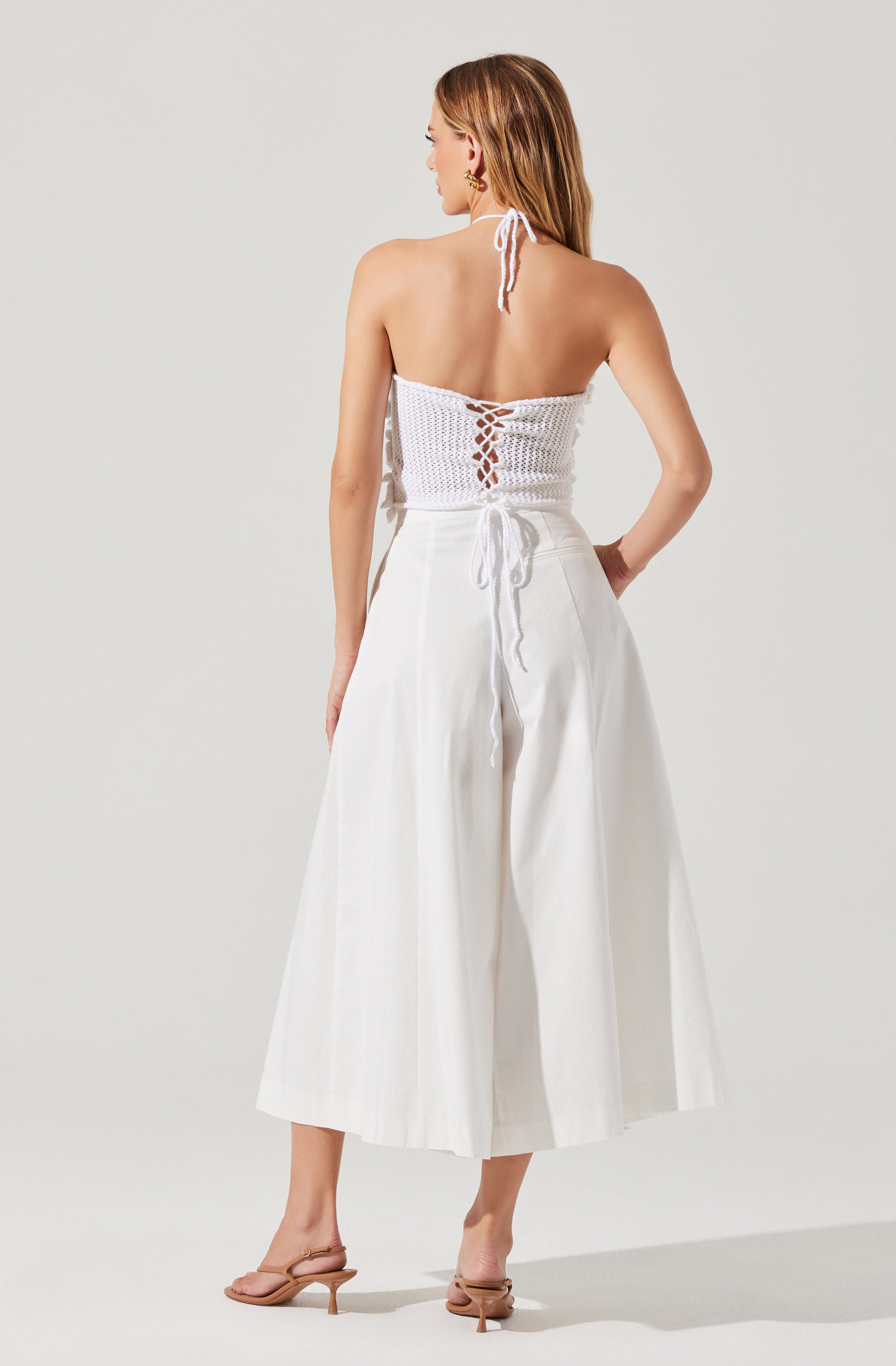 A woman is wearing a white knitted halter top with a lace-up back and a tie at the waist. She pairs it with high-waisted, wide-leg white trousers. She also has on beige heeled sandals and gold earrings. Her hair is styled down, and she is facing away from the camera.