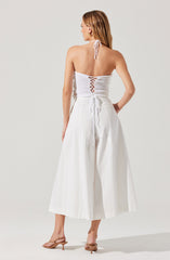 A woman is wearing a white knitted halter top with a lace-up back and a tie at the waist. She pairs it with high-waisted, wide-leg white trousers. She also has on beige heeled sandals and gold earrings. Her hair is styled down, and she is facing away from the camera.