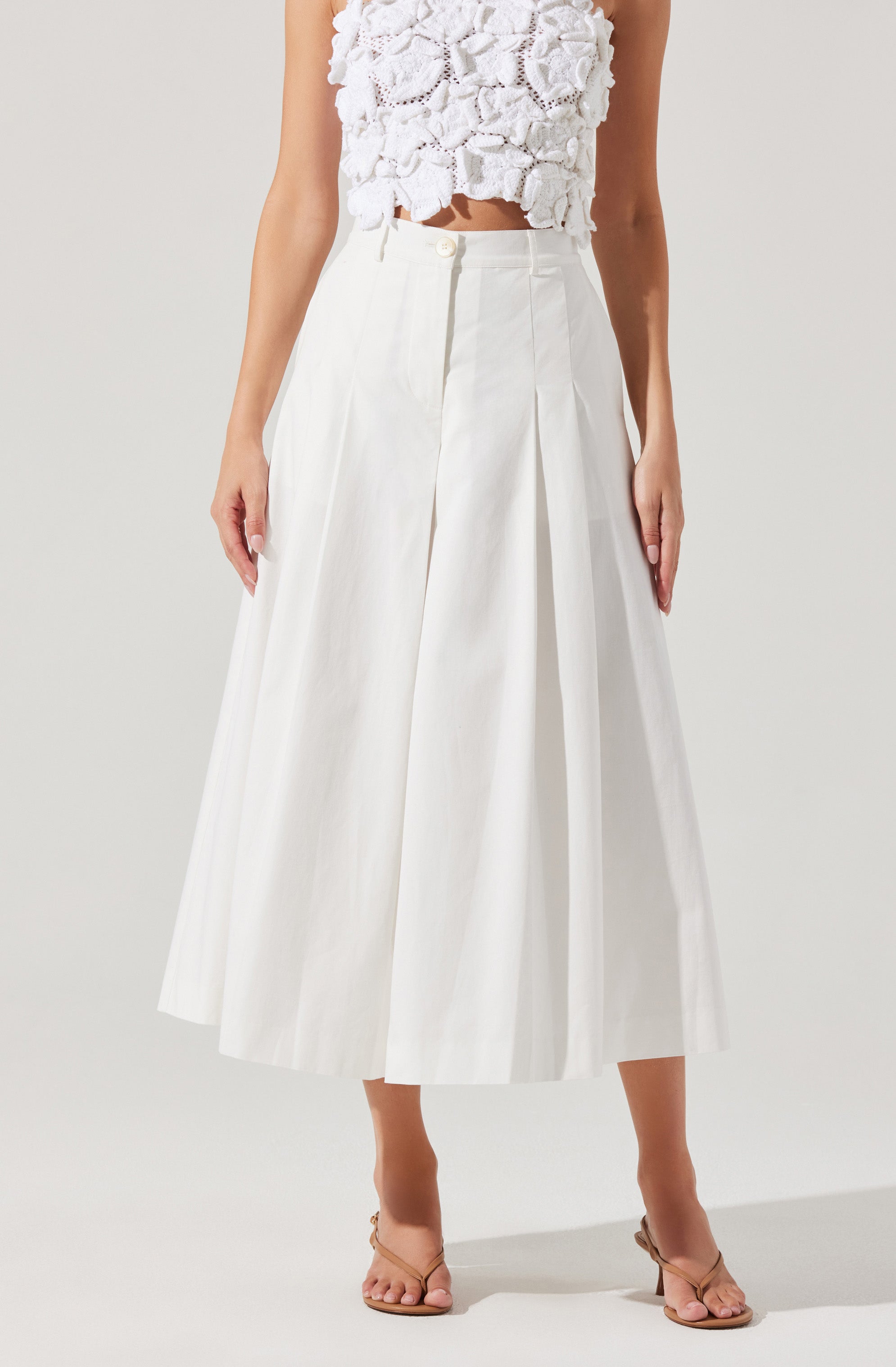 A woman is wearing a white sleeveless top with textured, floral fabric details. She is also wearing a high-waisted, white, wide-leg skirt with pleats, reaching mid-calf, and beige open-toe sandals.