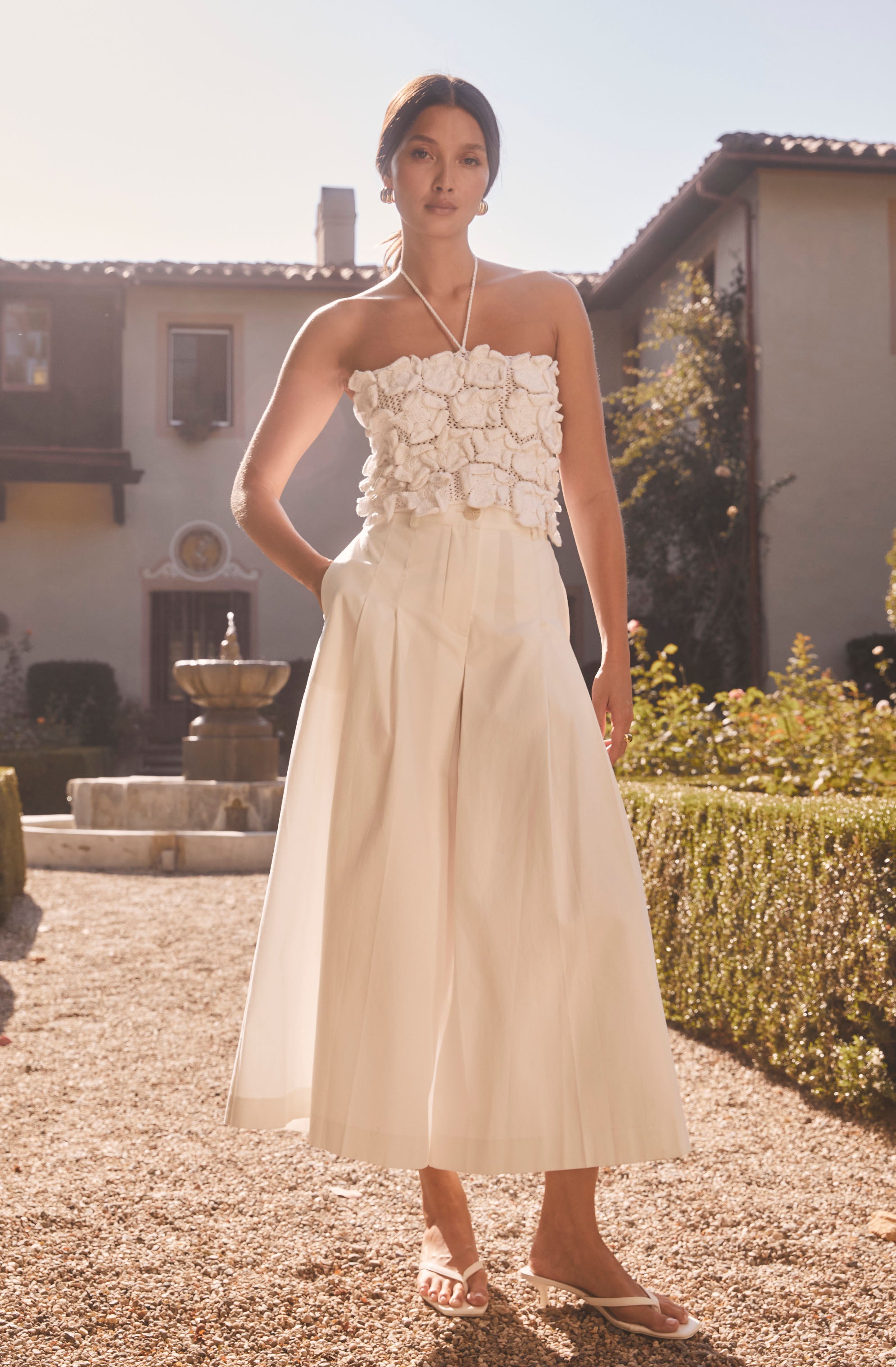 A woman is wearing a strapless top embellished with three-dimensional fabric flowers and a pair of high-waisted, wide-leg cream-colored trousers that reach her mid-calf. She is also wearing white sandals and has a simple necklace around her neck. The outfit has a light, elegant, and summery appearance.