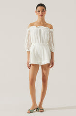 A woman is wearing an off-the-shoulder white blouse with ruffled edges and long, semi-sheer, eyelet sleeves. She pairs it with matching high-waisted white shorts that have a subtle eyelet pattern. She completes the outfit with open-toe, low-heeled sandals in a light, neutral color.