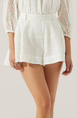 A person is wearing a matching set of white, eyelet lace clothing. The top has long sleeves and is semi-sheer with a floral cut-out pattern, with the fabric gathered at the waist. The shorts are high-waisted, made of the same eyelet lace material, with a wide waistband and a relaxed fit.