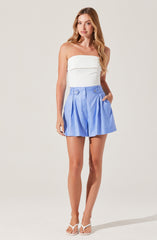 A woman is wearing a strapless white top with a wide horizontal band across the chest, paired with high-waisted light blue shorts that feature pleats, belt loops, and button details at the waist. She is also wearing white heeled sandals.