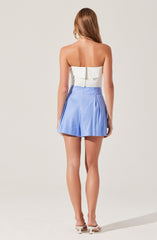 A woman wearing a strapless white top with a back zipper and light blue high-waisted shorts. The shorts feature pleats on the front and a slightly flared silhouette. She is also wearing white heeled shoes.