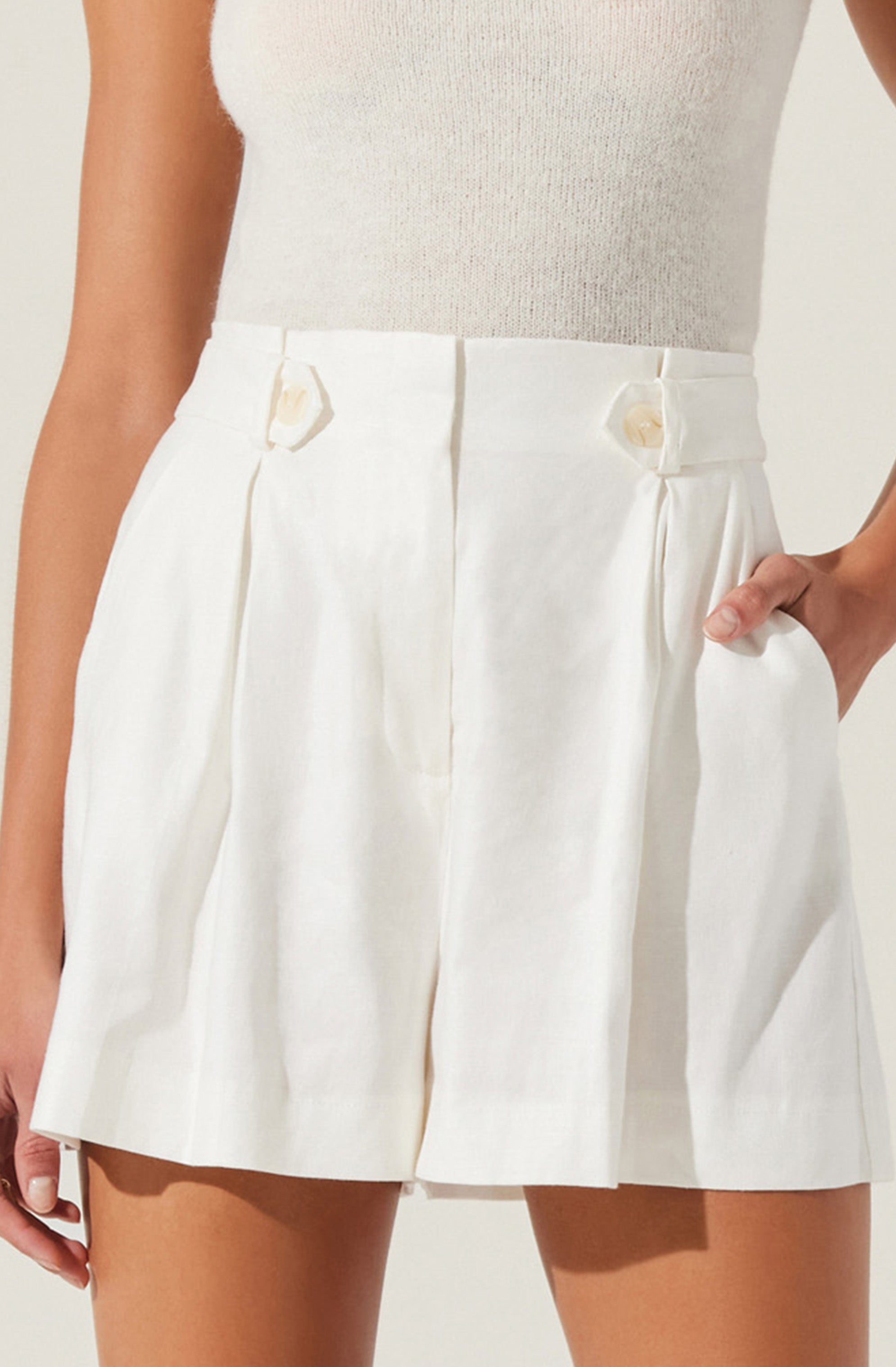 A person is wearing high-waisted white shorts with pleats and large round buttons at the waistband, paired with a sleeveless, light-colored knitted top. The shorts have a loose fit and side pockets.