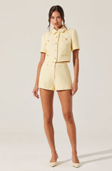 A woman is wearing a light yellow, textured, short-sleeved cropped jacket with a collar and decorative buttons on the front and pockets. She pairs it with matching high-waisted shorts made from the same textured fabric. She completes the outfit with light yellow pointed-toe heels.