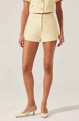A woman is wearing a light yellow, textured two-piece outfit consisting of a short-sleeved, button-up jacket and matching high-waisted shorts. She is also wearing beige pointed-toe high heels with ankle straps.