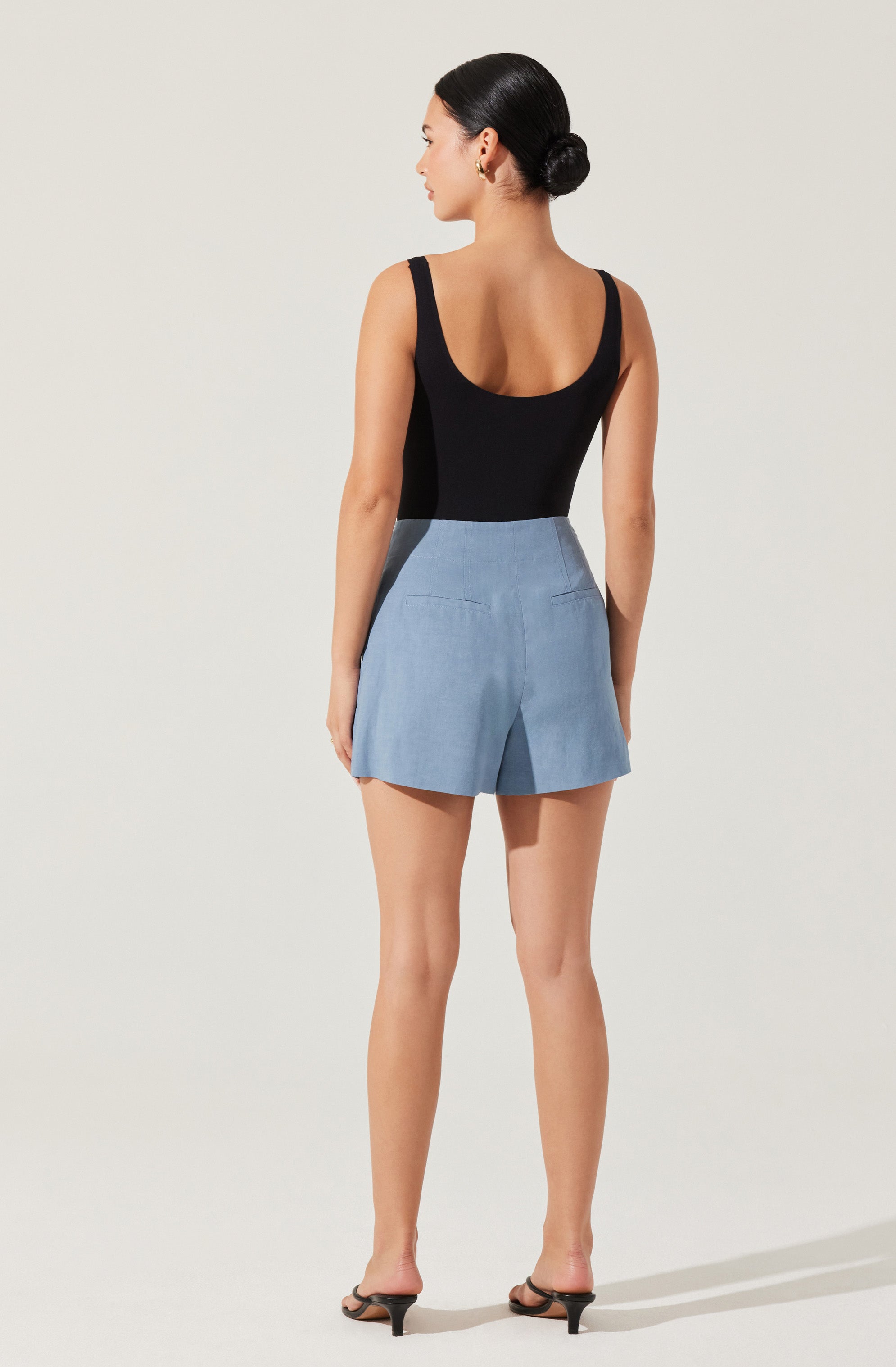 A woman is wearing a sleeveless black top with wide straps and a scoop neckline, paired with high-waisted light blue shorts that have a tailored fit and two back pockets. She is also wearing black open-toe heels with a thin strap across the toes and a small heel. Her hair is styled in a neat, low bun.