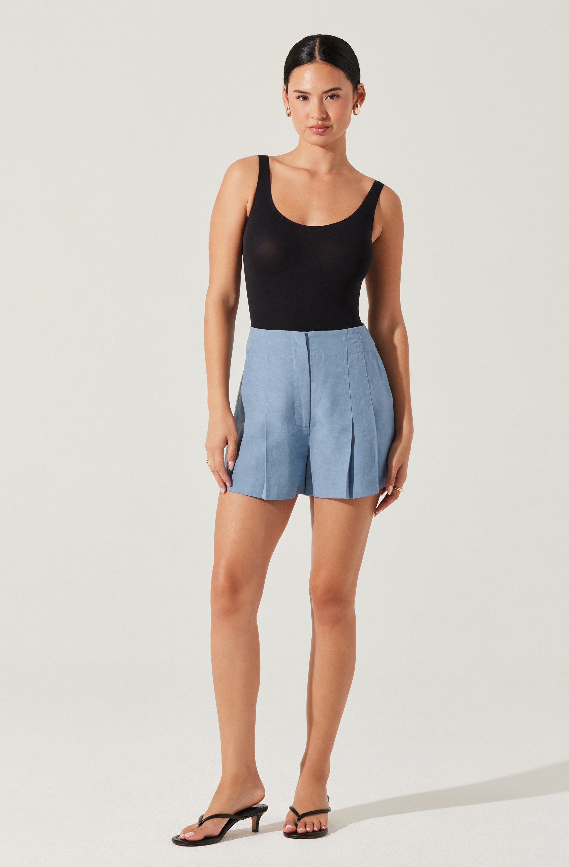 A woman is wearing a black sleeveless tank top with a scoop neckline, paired with high-waisted light blue pleated shorts. She has on black open-toe high-heeled sandals and small hoop earrings.