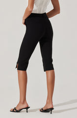 A woman is wearing black cropped pants with a high waist and a small slit at the hem on each leg. She is also wearing a sleeveless, light-colored top with a textured pattern. She has on black open-toe heeled sandals.