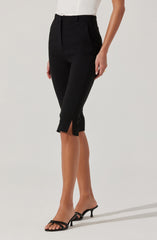 A person is wearing high-waisted black cropped pants with a small slit at the hem on each leg. The pants have a tailored fit with a waistband and belt loops. They are paired with an off-white or cream-colored blouse and black open-toe high-heeled sandals.