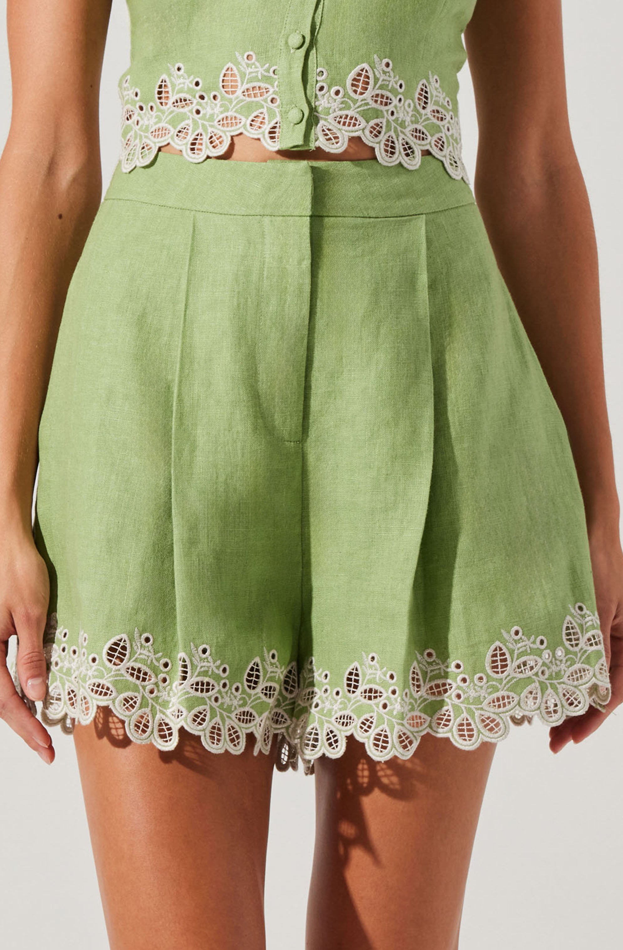 A woman is wearing a light green sleeveless top with decorative white floral embroidery along the hem and button area. The matching high-waisted shorts are also light green with the same white embroidered floral design along the hem, featuring intricate patterns of leaves and flowers.