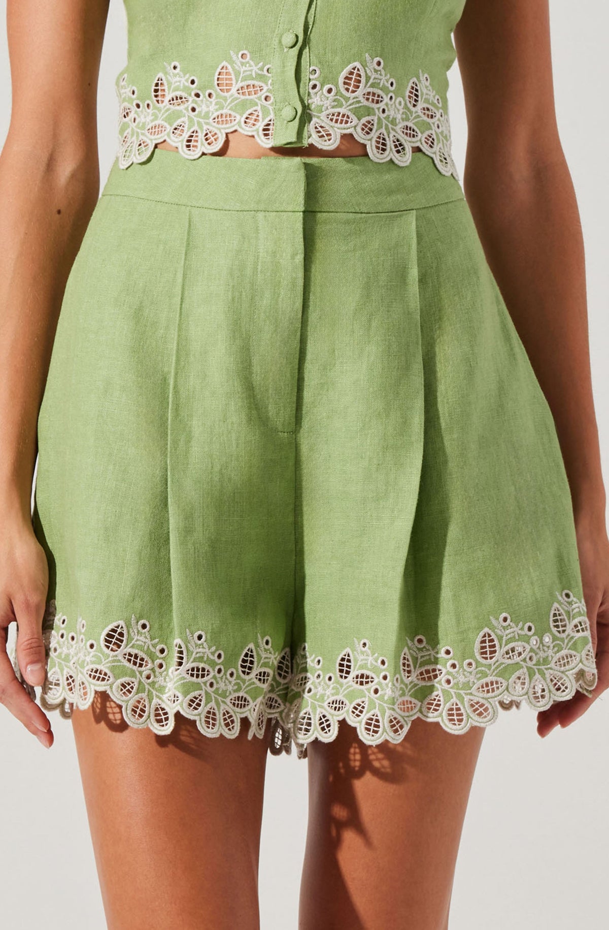 A woman is wearing a light green sleeveless top with decorative white floral embroidery along the hem and button area. The matching high-waisted shorts are also light green with the same white embroidered floral design along the hem, featuring intricate patterns of leaves and flowers.