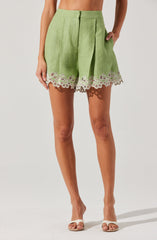 A person is wearing high-waisted, light green shorts with a pleated front and a wide waistband. The shorts feature decorative white lace trim with floral embroidery along the hem. They are paired with open-toe, beige sandals. The person's right hand is resting on their hip and their left hand is hanging down by their side.