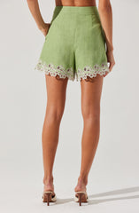 Light green shorts with a high waist and side pockets. The hem features white eyelet embroidery with floral and leaf patterns, giving a delicate and decorative touch. The shorts are paired with beige high-heeled shoes.
