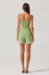 A woman is wearing a green sleeveless top with spaghetti straps tied into bows on her shoulders. The top is smocked and fitted at the waist. She is also wearing matching green shorts with white eyelet lace trim at the hem. The outfit appears light and summery. She is standing with her back to the camera, and her hair is down.