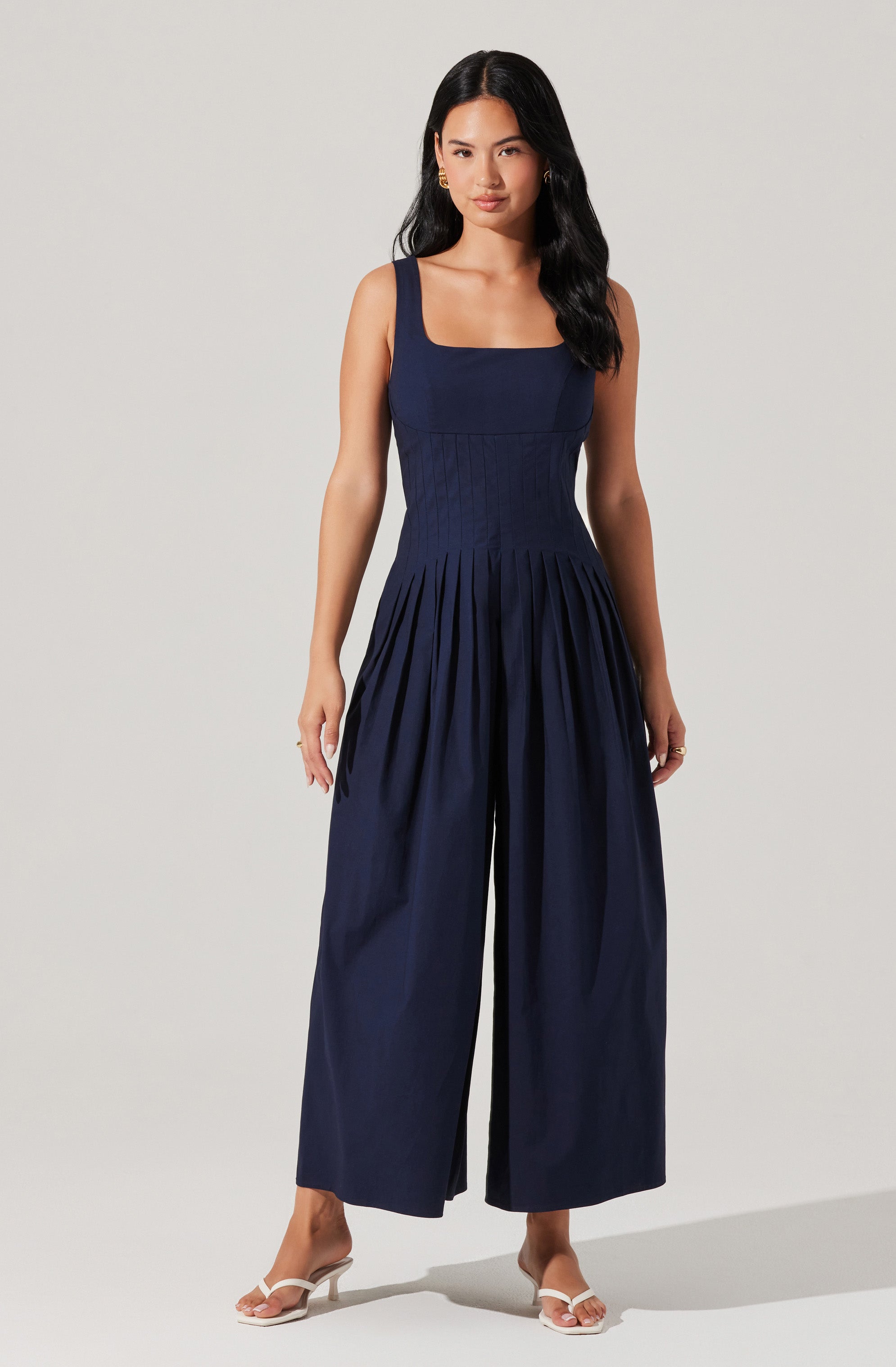 A woman is wearing a sleeveless navy blue jumpsuit with a square neckline. The fitted bodice features vertical seam details, and the wide-leg pants have pleats starting at the waist, creating a flowing silhouette. She is also wearing white heeled sandals.