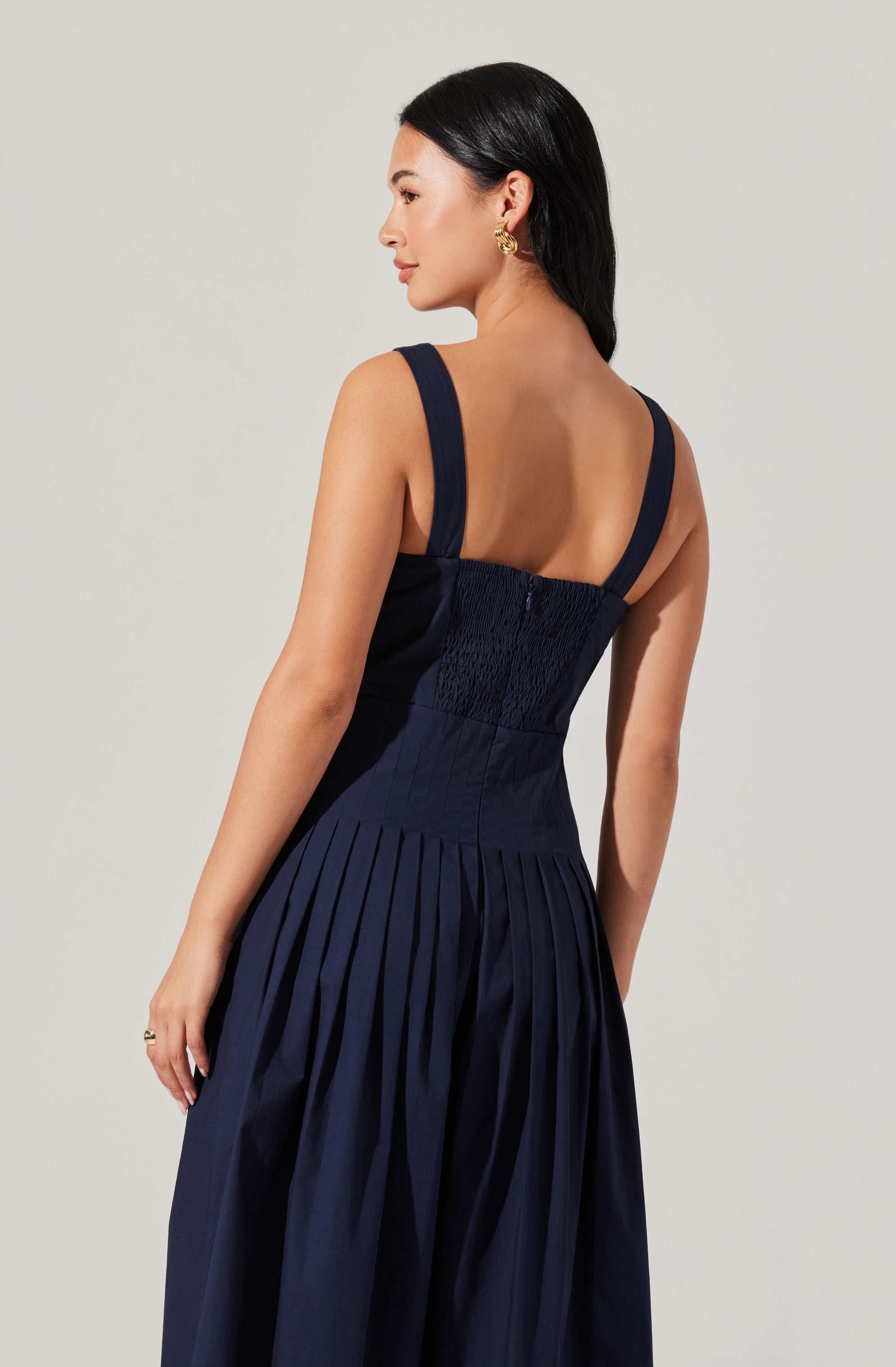 A woman is wearing a sleeveless navy blue dress with wide shoulder straps. The fitted bodice features smocking or shirring detail, and the skirt has evenly spaced, vertical pleats down its length.