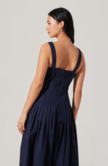 A woman is wearing a sleeveless navy blue dress with wide shoulder straps. The fitted bodice features smocking or shirring detail, and the skirt has evenly spaced, vertical pleats down its length.