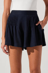 A person is wearing a pair of navy blue shorts with a high elastic waistband. The shorts feature vertical pleats or gathers that add texture, and they have a loose, flowy fit. The individual is also wearing a plain white sleeveless top.