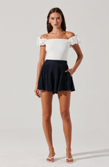 The woman is wearing an off-the-shoulder white top with puffed sleeves that have a textured, ruffled appearance. She pairs it with a high-waisted, navy blue skirt that is pleated and has a flared, ruffled hem. She completes the outfit with open-toed, beige high-heeled sandals.