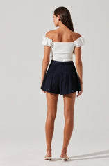 A woman is wearing an off-the-shoulder white top with ruffled sleeves and a fitted bodice. She pairs it with high-waisted, navy blue pleated shorts that have a wide waistband. She is also wearing white heeled shoes.