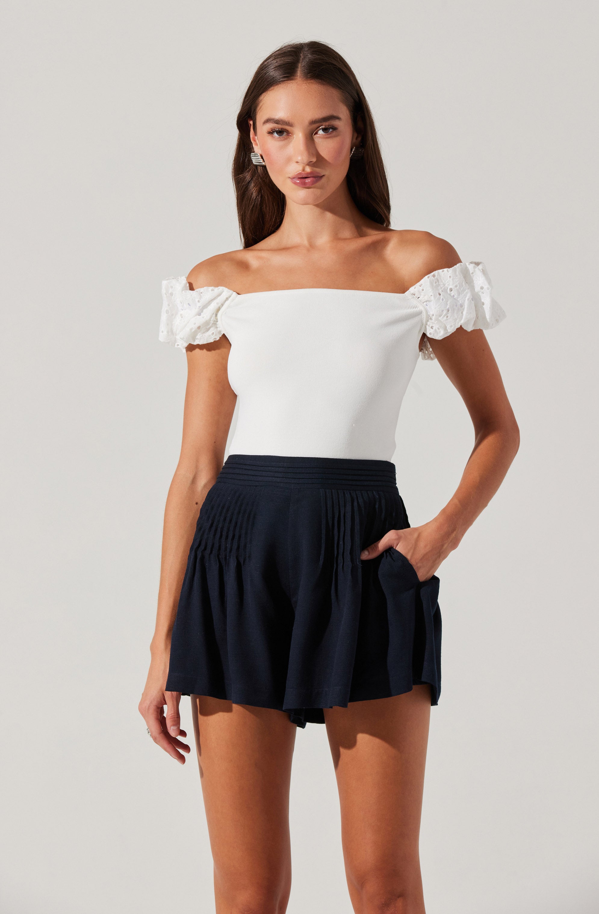 The woman is wearing an off-the-shoulder white top with ruffled lace sleeves and a pair of high-waisted black shorts with a gathered, pleated design.