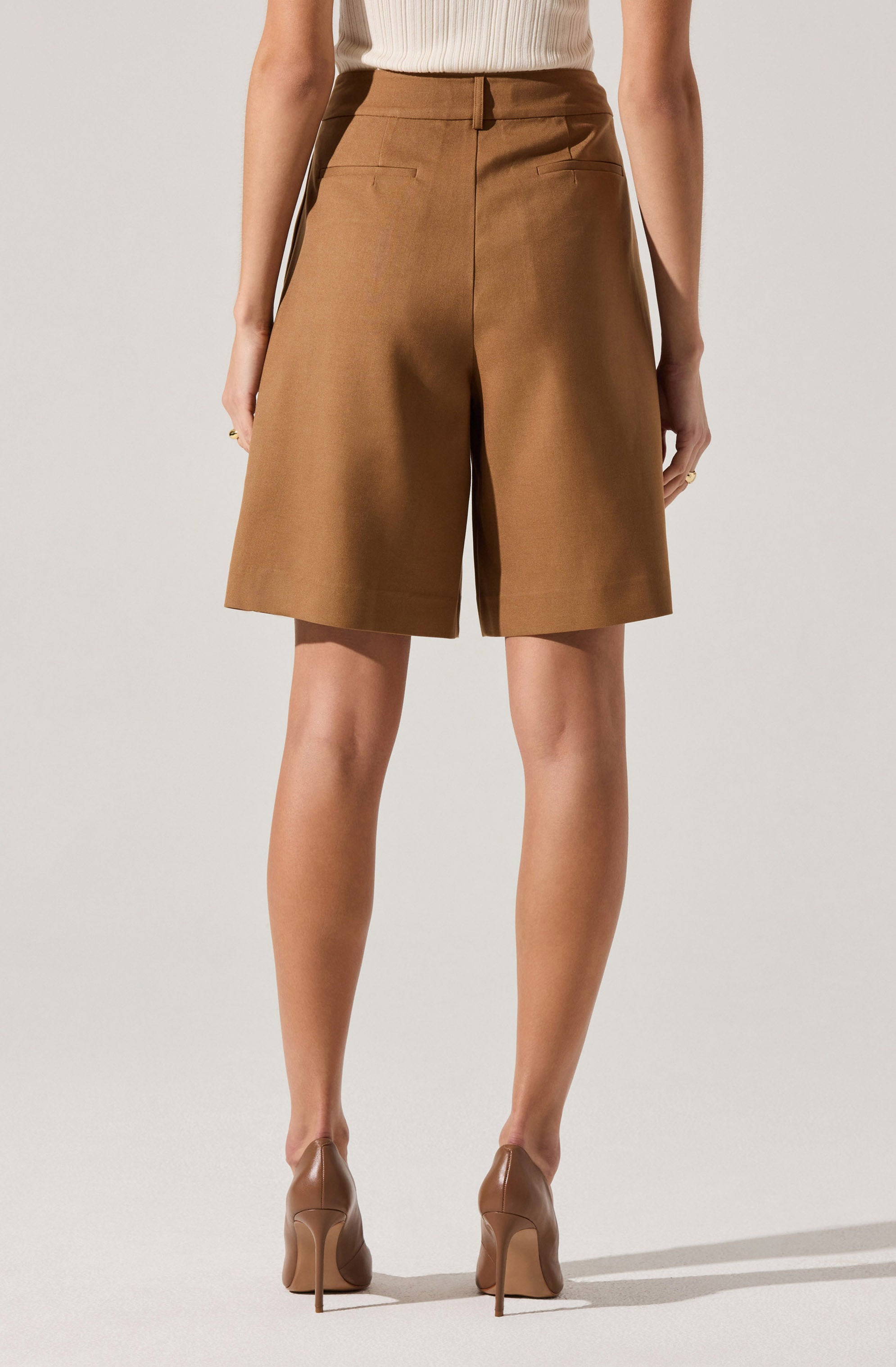 The image shows a person wearing high-waisted, knee-length brown shorts with two back pockets. They are paired with a sleeveless ribbed top in a light neutral color and beige high-heeled shoes.