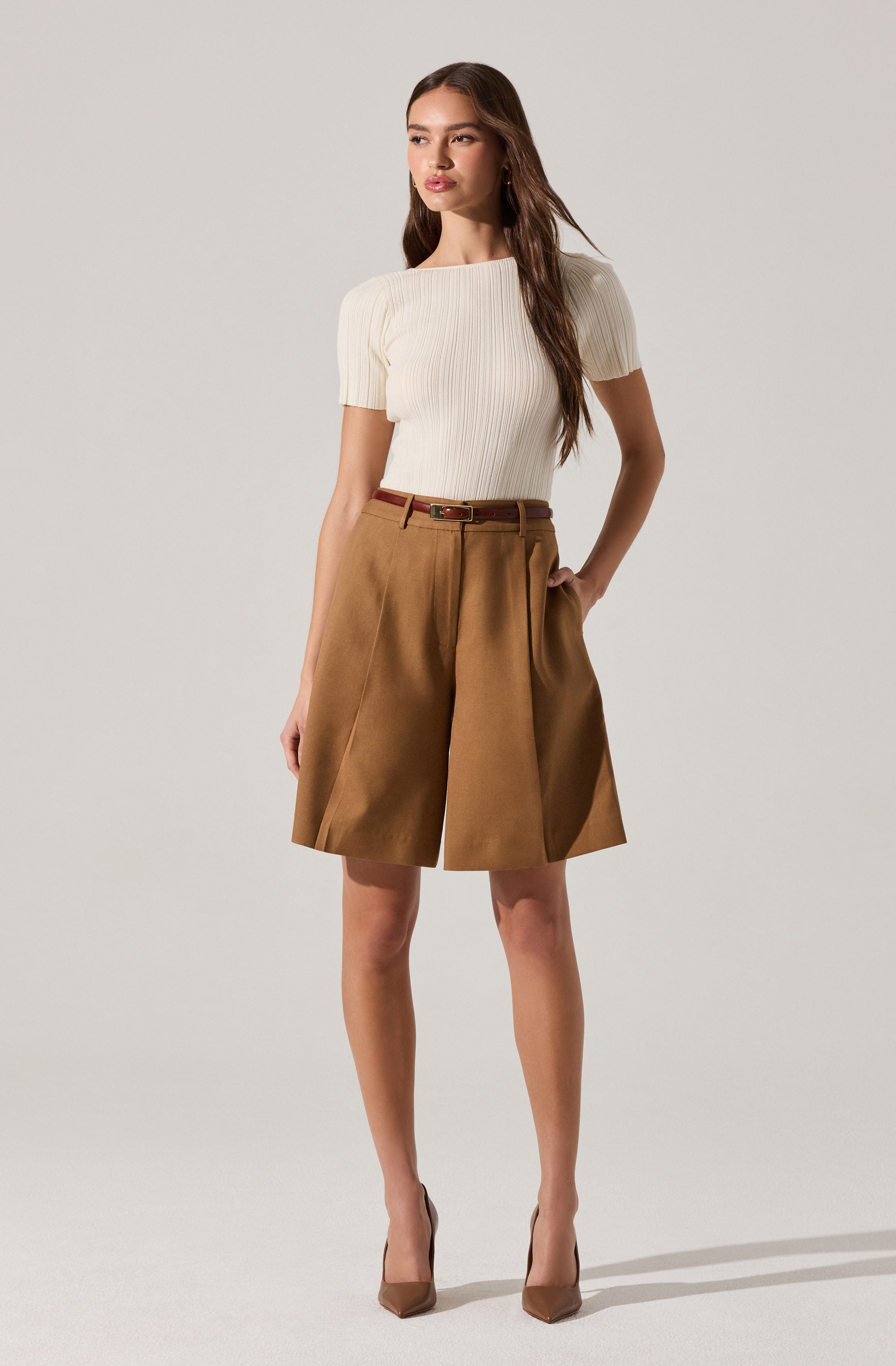 A woman is wearing a cream-colored, ribbed short-sleeve top with a boat neckline, tucked into high-waisted, tan knee-length shorts with pleats on the front. She is accessorized with a thin brown belt and is wearing nude-colored pointed-toe high heels.