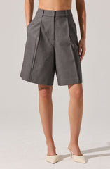 Gray tailored high-waisted shorts with wide pleats, belt loops, and front pleats. The shorts have a structured, slightly loose fit and feature sharp creases down the front. The model is wearing cream-colored pointed-toe high heels.