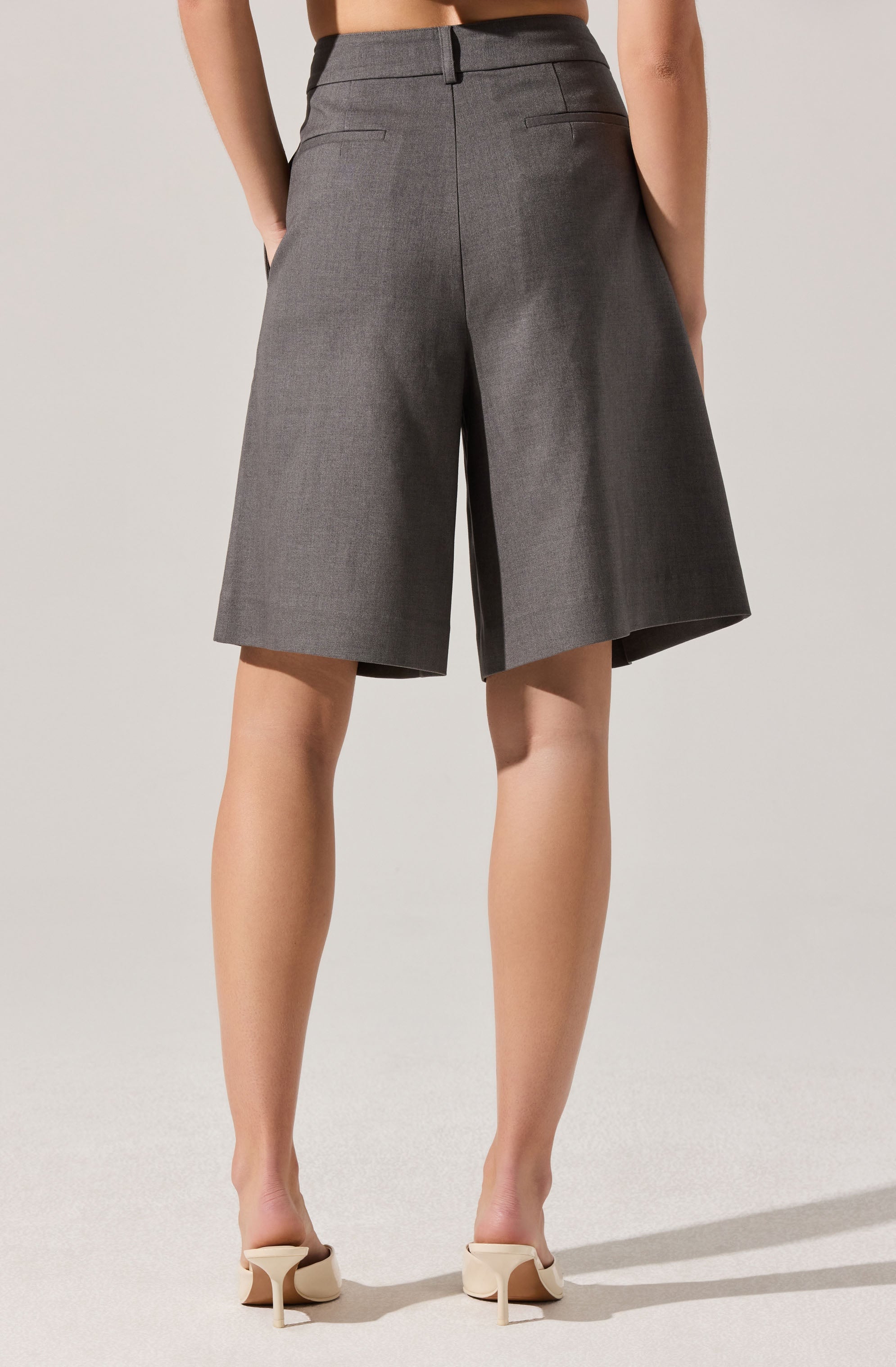 A person is wearing high-waisted gray shorts made of a textured fabric with a tailored fit. The shorts have a waistband with belt loops, a center back seam, and two horizontal slit pockets at the back. The hem falls just above the knees. The person is also wearing beige high-heeled shoes with a pointed toe and slim heel.